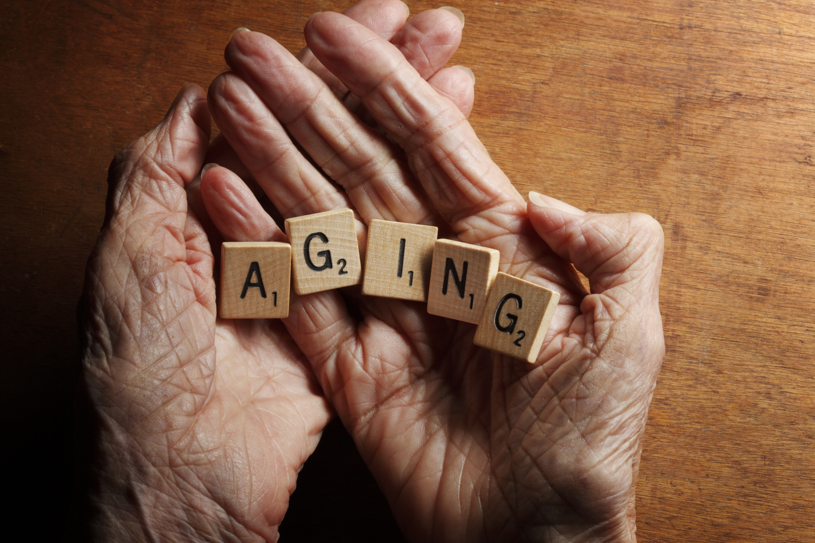 Twee oudere dames handen met de handpalmen open op een houten tafel. In de handpalm liggen scrabble steentjes die het woord: aging vormen.