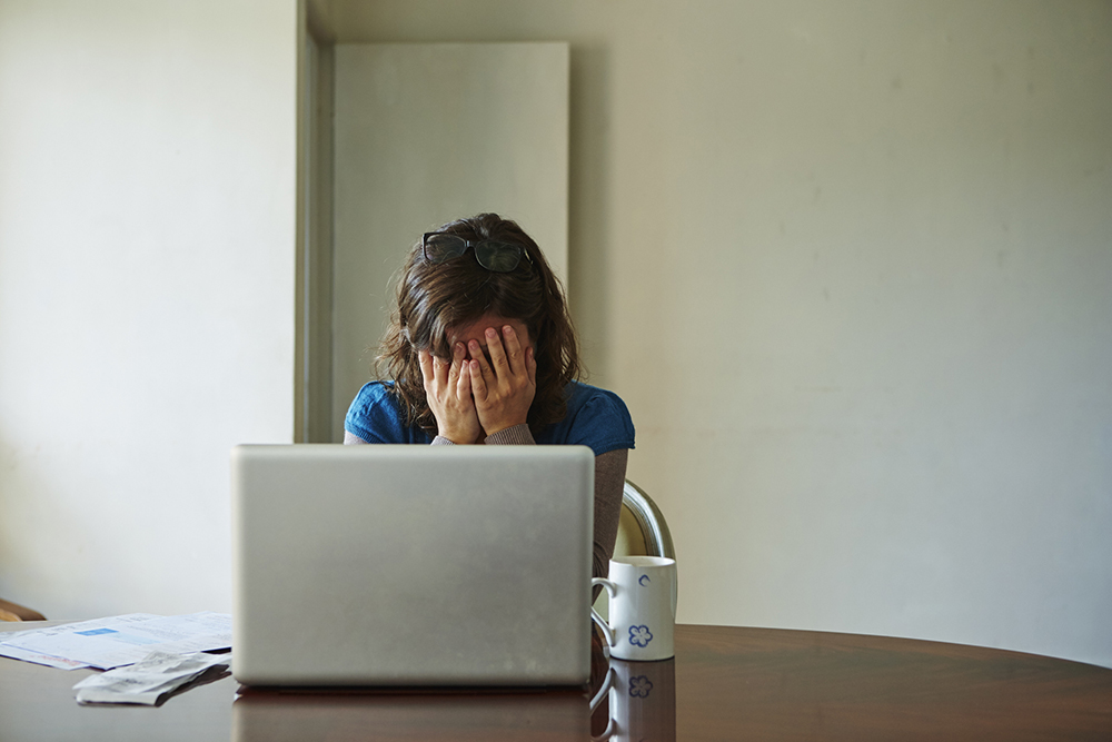 Vrouw met geldzorgen houdt haar handen voor haar gezicht terwijl ze aan tafel achter haar laptop zit.