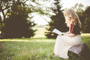 Een blonde vrouw op een steen midden op een grasveld leest een boek
