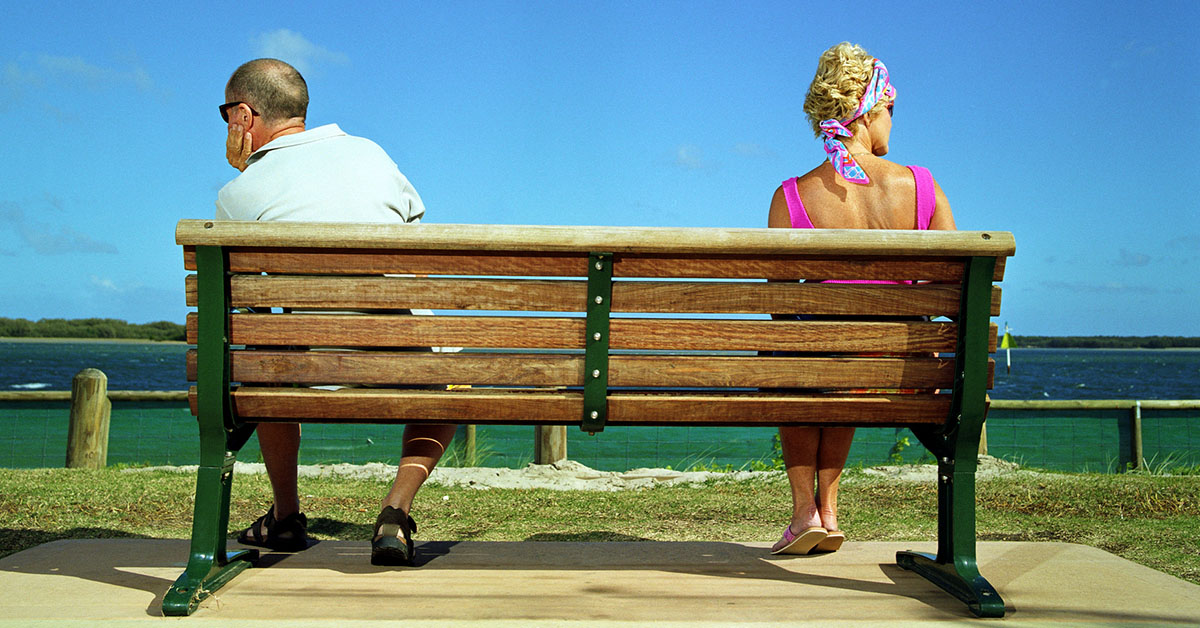 Man en vrouw zitten elk aan een kant van een bankje en kijken uit op zee